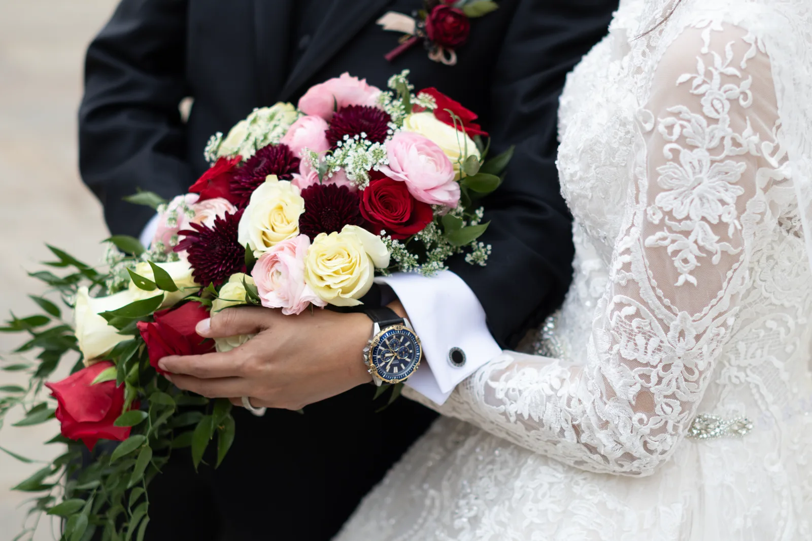 Close-up of a bride and groom holding a bouquet of red, cream, and burgundy roses